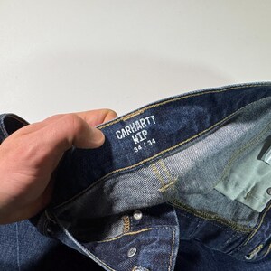 Puede incluir: Primer plano de unos vaqueros de mezclilla azul Carhartt WIP. Los vaqueros tienen bragueta con botones y la etiqueta dice "CARHARTT WIP 34/34". La mezclilla es un lavado azul oscuro con costuras y remaches met&aacute;licos visibles. Los vaqueros est&aacute;n doblados.