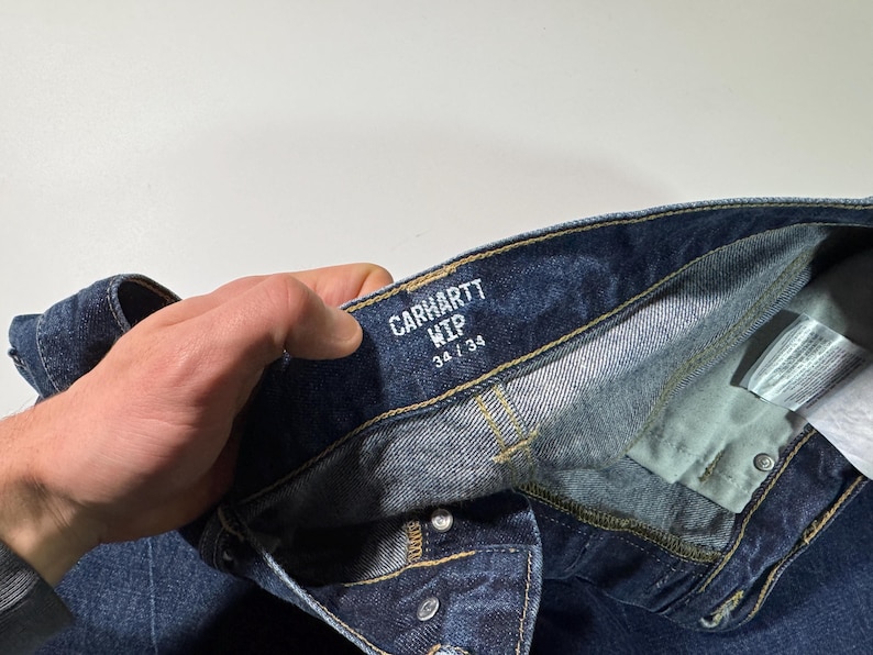 Puede incluir: Primer plano de unos vaqueros de mezclilla azul Carhartt WIP. Los vaqueros tienen bragueta con botones y la etiqueta dice "CARHARTT WIP 34/34". La mezclilla es un lavado azul oscuro con costuras y remaches met&aacute;licos visibles. Los vaqueros est&aacute;n doblados.