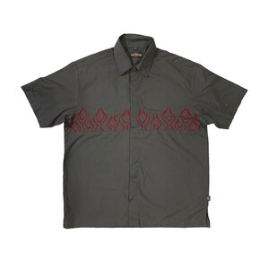 Puede incluir: Una camisa de manga corta de color gris oscuro con botones y bordado de llamas rojas en el pecho. La camisa tiene un cuello abotonado y un solo bolsillo en el lado izquierdo.