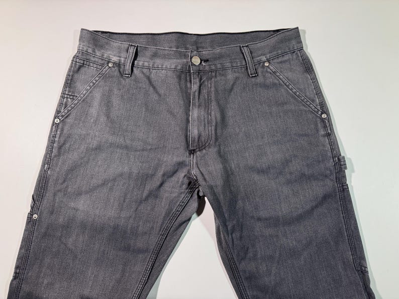 Peut inclure: Pantalon de travail en denim gris avec plusieurs poches et coutures renforc&eacute;es. Le pantalon a une fermeture boutonn&eacute;e et des rivets aux poches. Le tissu semble pr&eacute;-lav&eacute;. Un style de v&ecirc;tement de travail classique.