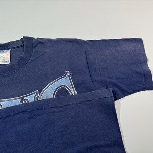 May include: A navy blue t-shirt with a light blue and white graphic design. The graphic design features a stylized logo with a starburst effect.