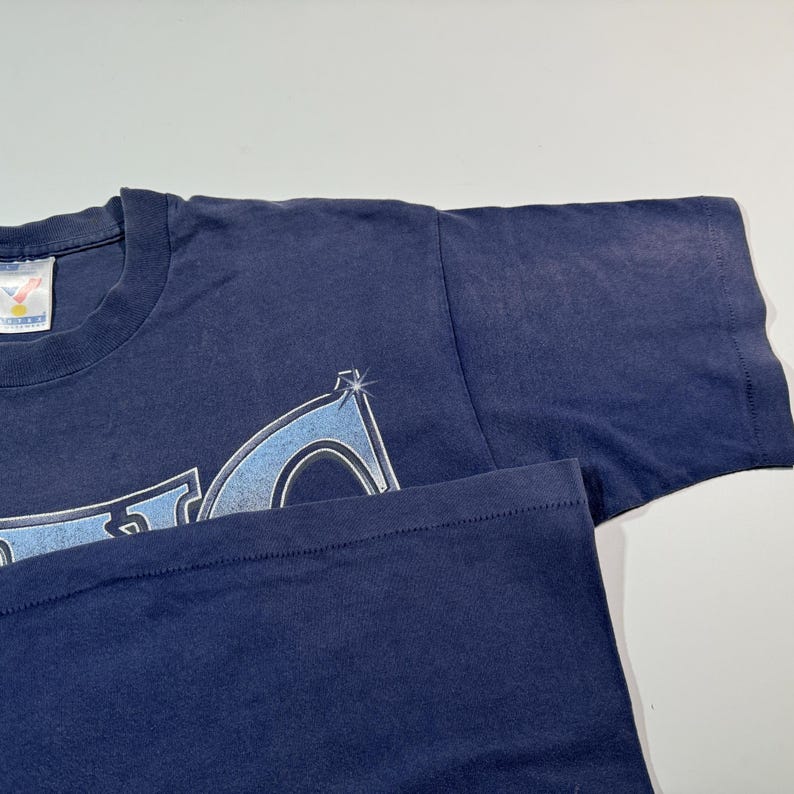 May include: A navy blue t-shirt with a light blue and white graphic design. The graphic design features a stylized logo with a starburst effect.