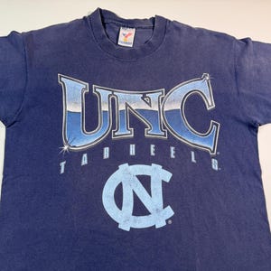 May include: A navy blue t-shirt with a vintage UNC Tar Heels graphic. The graphic features the letters "UNC" in a blue and white gradient with a silver outline. The word "Tar Heels" is written in white below the letters. A blue and white UNC logo is centered below the text.