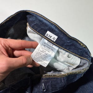 Puede incluir: Primer plano de unos vaqueros de mezclilla azul oscuro. Los vaqueros tienen una etiqueta visible con el logotipo de Carhartt e informaci&oacute;n del producto. Los vaqueros est&aacute;n doblados, mostrando el forro interior y los detalles de las costuras. Tambi&eacute;n es visible una peque&ntilde;a etiqueta blanca con la fecha "10.25.12".