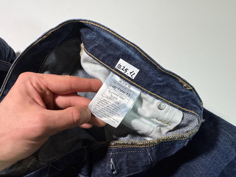 Puede incluir: Primer plano de unos vaqueros de mezclilla azul oscuro. Los vaqueros tienen una etiqueta visible con el logotipo de Carhartt e informaci&oacute;n del producto. Los vaqueros est&aacute;n doblados, mostrando el forro interior y los detalles de las costuras. Tambi&eacute;n es visible una peque&ntilde;a etiqueta blanca con la fecha "10.25.12".