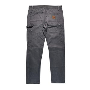 Peut inclure: Pantalon de travail gris avec plusieurs poches et un patch en cuir marron. Le pantalon a un design classique et durable, adapt&eacute; &agrave; diverses t&acirc;ches. Le logo Carhartt est visible sur la poche arri&egrave;re.