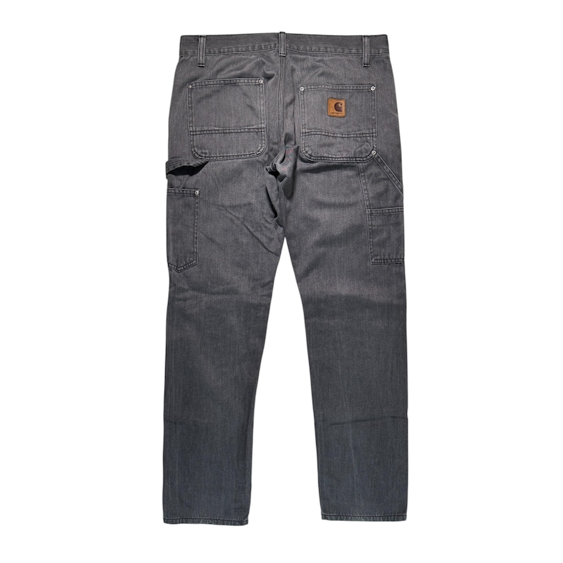 Peut inclure: Pantalon de travail gris avec plusieurs poches et un patch en cuir marron. Le pantalon a un design classique et durable, adapt&eacute; &agrave; diverses t&acirc;ches. Le logo Carhartt est visible sur la poche arri&egrave;re.