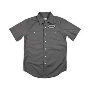 Puede incluir: Una camisa gris de manga corta con botones y un parche con el logotipo de Carhartt en el pecho izquierdo.