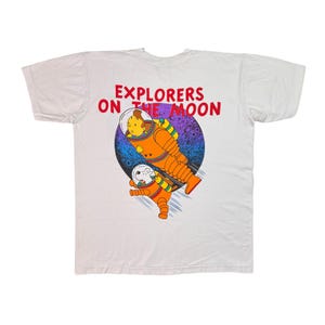 May include: A white short-sleeve t-shirt featuring a graphic of two figures in orange space suits floating in front of a dark blue moon. The text EXPLORERS ON THE MOON is printed in red above the graphic, which depicts a person and a dog.
