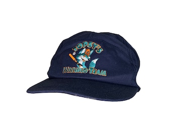 Puede incluir: Una gorra de béisbol azul marino con un logotipo bordado de un lobo de dibujos animados que lleva una gorra de béisbol y sostiene un bate. El logotipo dice "Topsy's Winning Team".