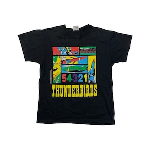 Puede incluir: Camiseta negra con un gráfico colorido de los Thunderbirds, un equipo de dibujos animados de astronautas y sus vehículos. El gráfico incluye los números 54321 y el texto "THUNDERBIRDS".