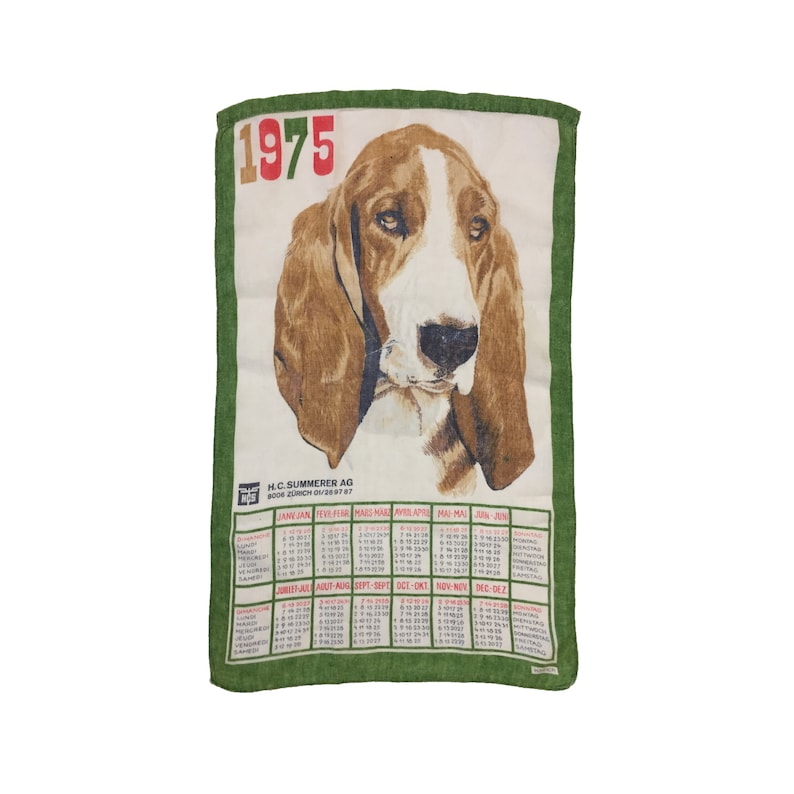 Puede incluir: Una toalla de cocina verde y blanca con un calendario para el a&ntilde;o 1975 y una ilustraci&oacute;n de un perro sabueso marr&oacute;n y blanco. El texto en la toalla dice "H.C. Summerer AG 8006 Zurich 01/29 87".