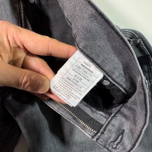 Peut inclure: Gros plan sur un jean Carhartt gris avec une &eacute;tiquette visible. L'&eacute;tiquette affiche le nom de la marque, les d&eacute;tails du style et la taille 32 x 32. Le jean a une fermeture &eacute;clair et un bouton.