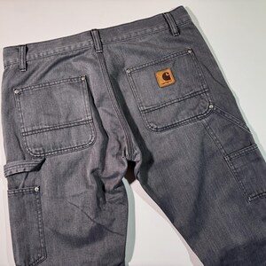 Peut inclure: Pantalon de travail gris avec plusieurs poches et un patch en cuir marron. Le pantalon a un design classique avec des coutures renforc&eacute;es et une construction durable. Le logo Carhartt est visible sur la poche arri&egrave;re.