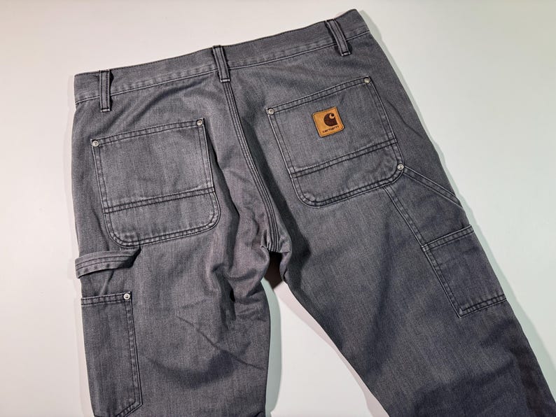 Peut inclure: Pantalon de travail gris avec plusieurs poches et un patch en cuir marron. Le pantalon a un design classique avec des coutures renforc&eacute;es et une construction durable. Le logo Carhartt est visible sur la poche arri&egrave;re.
