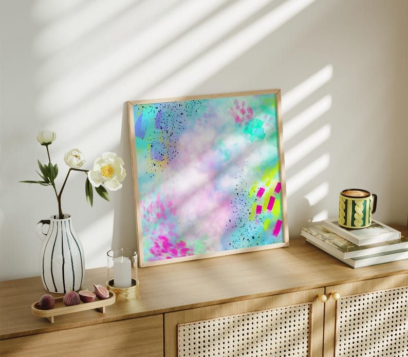 May include: A framed abstract painting with a light blue, pink, and yellow background. The painting has a splattered effect with black, pink, and green accents.