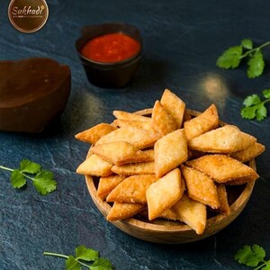 May include: A wooden bowl overflowing with golden-brown, diamond-shaped snacks. A small black bowl of red sauce is behind the snacks. Fresh green coriander leaves are scattered around. The Sukhadhi Kitchen logo is in the upper left corner.