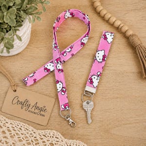 May include: A pink fabric lanyard and a matching wristlet keychain, both featuring a repeating Hello Kitty pattern with white faces and red bows. The wristlet includes a silver metal clasp holding a silver key. A brown paper tag reads Crafty Angie.