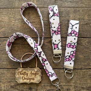 May include: A fabric lanyard and two matching wristlet keychains featuring a repeating Hello Kitty pattern with pink bows on a white background. Each item includes silver-toned metal hardware, including split rings and a lobster claw clasp.