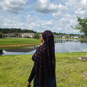 May include: A black headscarf featuring a repeating pattern of small, dark red, leaf-shaped motifs. The lightweight fabric drapes over the head and shoulders, showing a consistent print throughout the material, worn over a solid black long-sleeved top.