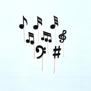 May include: Black glitter musical note and symbol cake toppers on wooden sticks. The symbols include a treble clef, a sharp, a flat, and a music note.