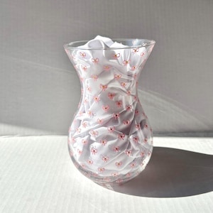 May include: A clear glass vase with a pink bow pattern. The vase is empty and has a wide mouth.