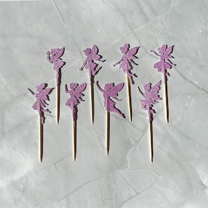 May include: Eight glittery purple fairy cupcake toppers. Each fairy is a silhouette with outstretched arms and wings, mounted on a wooden pick. The fairies are arranged on a light-coloured surface, ideal for a themed party.