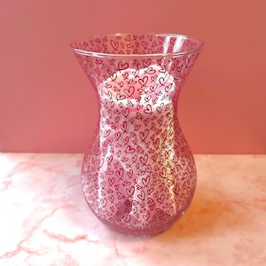 May include: A clear glass vase with a pink hue, decorated with a pattern of small, dark pink hearts. The vase has a flared top and a rounded base. A white candle is inside, creating a soft glow. The background is a solid pink colour.
