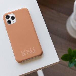 May include: A light brown phone case with a white camera lens and the letters "KNJ" printed on the back.