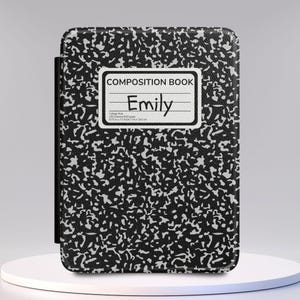 May include: A rectangular protective case for an electronic device featuring a classic black and white speckled composition book pattern. A white label on the front displays the text COMPOSITION BOOK and the name Emily in black handwritten script.