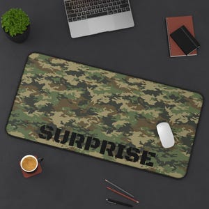 Puede incluir: Un tapete de escritorio de camuflaje verde y marrón con la palabra "SURPRISE" impresa en letras negras.