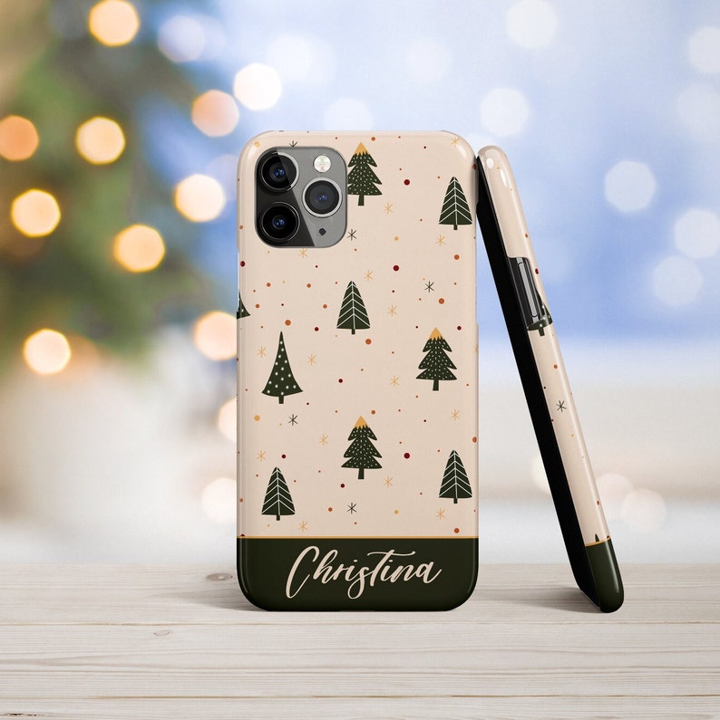 K&ouml;nnte beinhalten: Eine personalisierte Handyh&uuml;lle mit cremefarbenem Hintergrund und einem Muster aus gr&uuml;nen Weihnachtsb&auml;umen, roten und goldenen Punkten und wei&szlig;en Schneeflocken. Der Name "Christina" ist unten auf der H&uuml;lle in schwarzer Kursivschrift geschrieben.