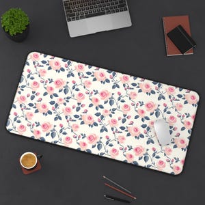 May include: A white desk mat with a pink and blue floral pattern. The mat is on a black surface with a laptop, a notebook, a cup of coffee, and a computer mouse.