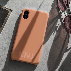 May include: A peach-colored phone case with the letters "KNJ" printed on the back.