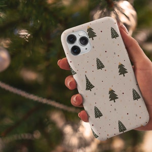 K&ouml;nnte beinhalten: Eine wei&szlig;e Handyh&uuml;lle mit einem Muster aus gr&uuml;nen Weihnachtsb&auml;umen und goldenen Sternen. Die H&uuml;lle wird in einer Hand gehalten.