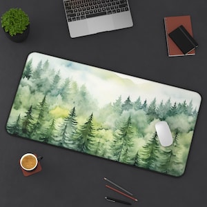 May include: A green and white watercolor forest scene printed on a rectangular mouse pad. The mouse pad is on a black surface with a laptop, a cup of coffee, pencils, and a notebook.