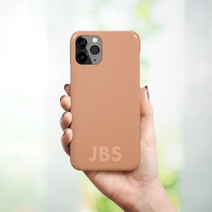 May include: A light brown phone case with a black camera lens and the initials "JBS" printed on the back.