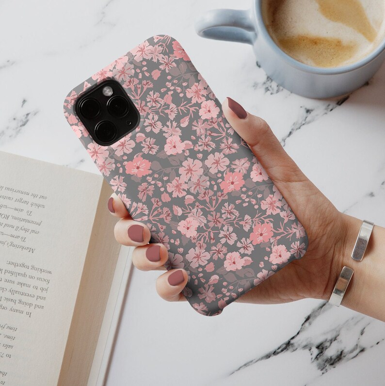 Puede incluir: Una funda de tel&eacute;fono gris con un patr&oacute;n floral rosa. La funda est&aacute; dise&ntilde;ada para un iPhone con un recorte para la c&aacute;mara.