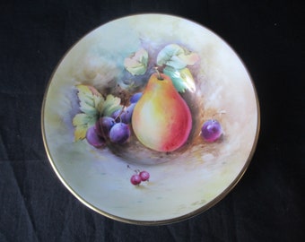 May include: A white ceramic bowl with a gold rim features a hand-painted design of a pear and grapes. The pear is yellow and red, and the grapes are purple. The bowl is on a black background.