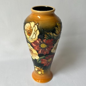 May include: A tall, decorative vase with a flared shape. The vase features a floral design with cream, red, and yellow flowers against a dark background. The vase has a glossy finish and a warm, orange-brown color.