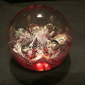 May include: A clear glass paperweight with a red and white swirl design. The paperweight has several air bubbles trapped inside.