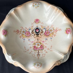 May include: An antique, scalloped-edge serving dish with a cream-colored base. The dish features a floral design in pink, blue, yellow, and white, with gold trim. The center of the dish has a detailed floral arrangement, and the edges are gently curved.