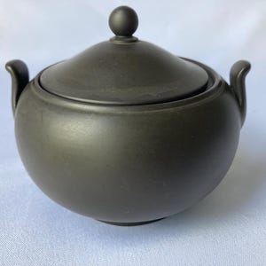 May include: A dark gray ceramic sugar bowl with a matching lid. The bowl has a rounded shape with two small handles and a lid with a small, round knob. The sugar bowl is a matte finish and sits on a white surface.