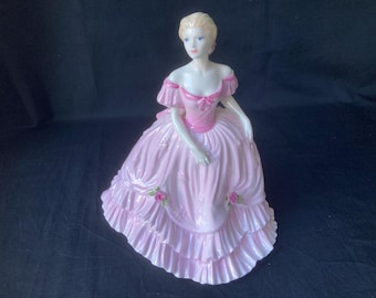May include: A porcelain figurine of a woman wearing a pink dress with ruffles and floral accents. The figurine is posed in a seated position with her hands resting on her lap.