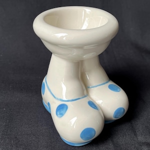Puede incluir: Un divertido huevero de cerámica blanca con forma de cuenco y dos pies. Los pies están decorados con lunares azules y un ribete azul. El huevero está diseñado para sostener un solo huevo.