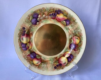 May include: A white ceramic plate with a gold rim and a fruit design. The plate features a variety of fruits, including plums, pears, and apples, painted in shades of purple, red, and green. The center of the plate is a gold-colored circle.