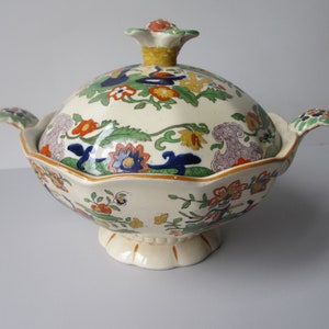 May include: A white ceramic serving dish with a lid. The dish features a floral pattern in shades of blue, green, yellow, orange, and red. The lid has a knob in the shape of a flower.