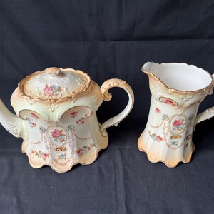 May include: A vintage porcelain tea set, featuring a teapot with a lid and a creamer. The set is decorated with floral patterns in pink, blue, and peach, with gold trim. The teapot has a curved spout and handle, and the creamer has a handle and a flared rim.