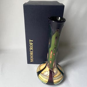 May include: A tall, slender ceramic vase with a flared rim, featuring a dark purple, green, and cream-colored pattern. Behind the vase stands a dark blue rectangular box with the word MOORCROFT printed in gold lettering on the front.