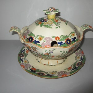 May include: A vintage cream-coloured porcelain gravy boat with a lid and a matching plate. The boat and plate are decorated with a colourful floral pattern in shades of blue, green, orange, and yellow.